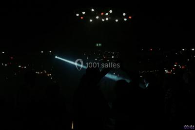 Homme souriant avec une moustache, portant une chemise sombre, devant un arrière-plan flou de verdure.
