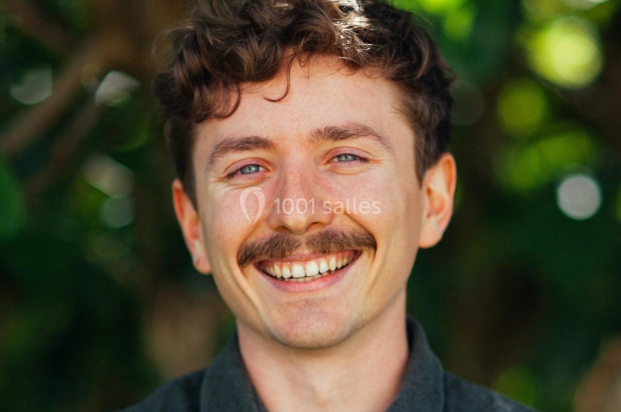Homme souriant avec une moustache, portant une chemise sombre, devant un arrière-plan flou de verdure.
