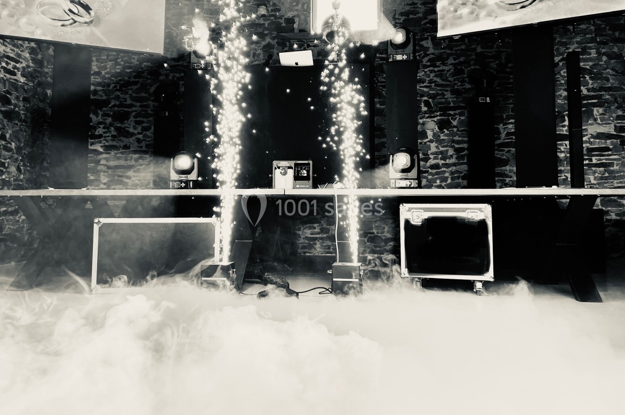 Scène de concert avec jeux de lumière, fumée au sol et équipements audio devant un mur en pierre.