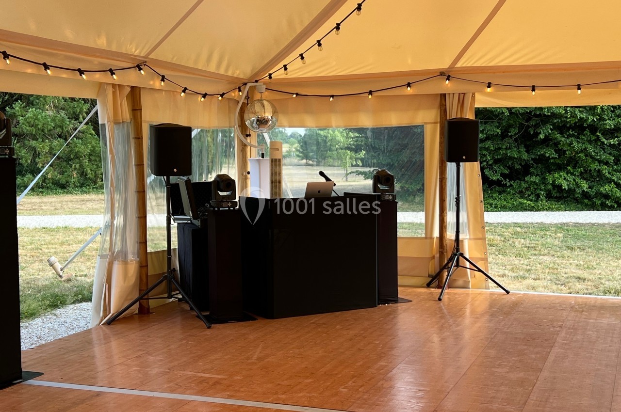 Stand de DJ sous une tente avec éclairage suspendu, piste en bois et environnement extérieur verdoyant.