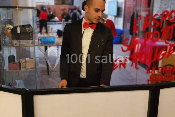 Un homme en costume noir et nœud papillon rouge se tient derrière un comptoir dans un espace intérieur.