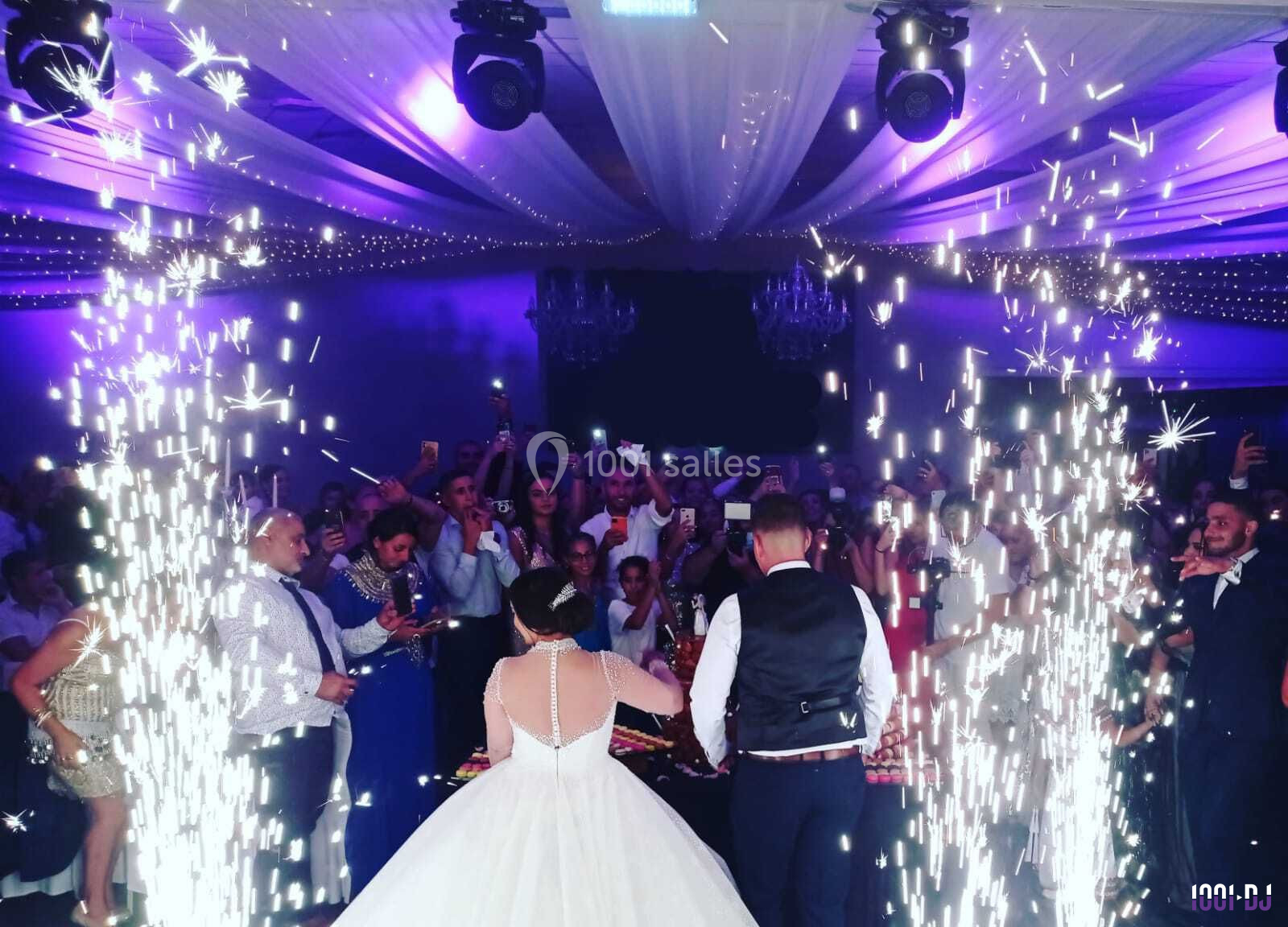 Un couple de mariés coupe leur gâteau sous des étincelles, entouré d'invités dans une salle décorée de lumières violettes.