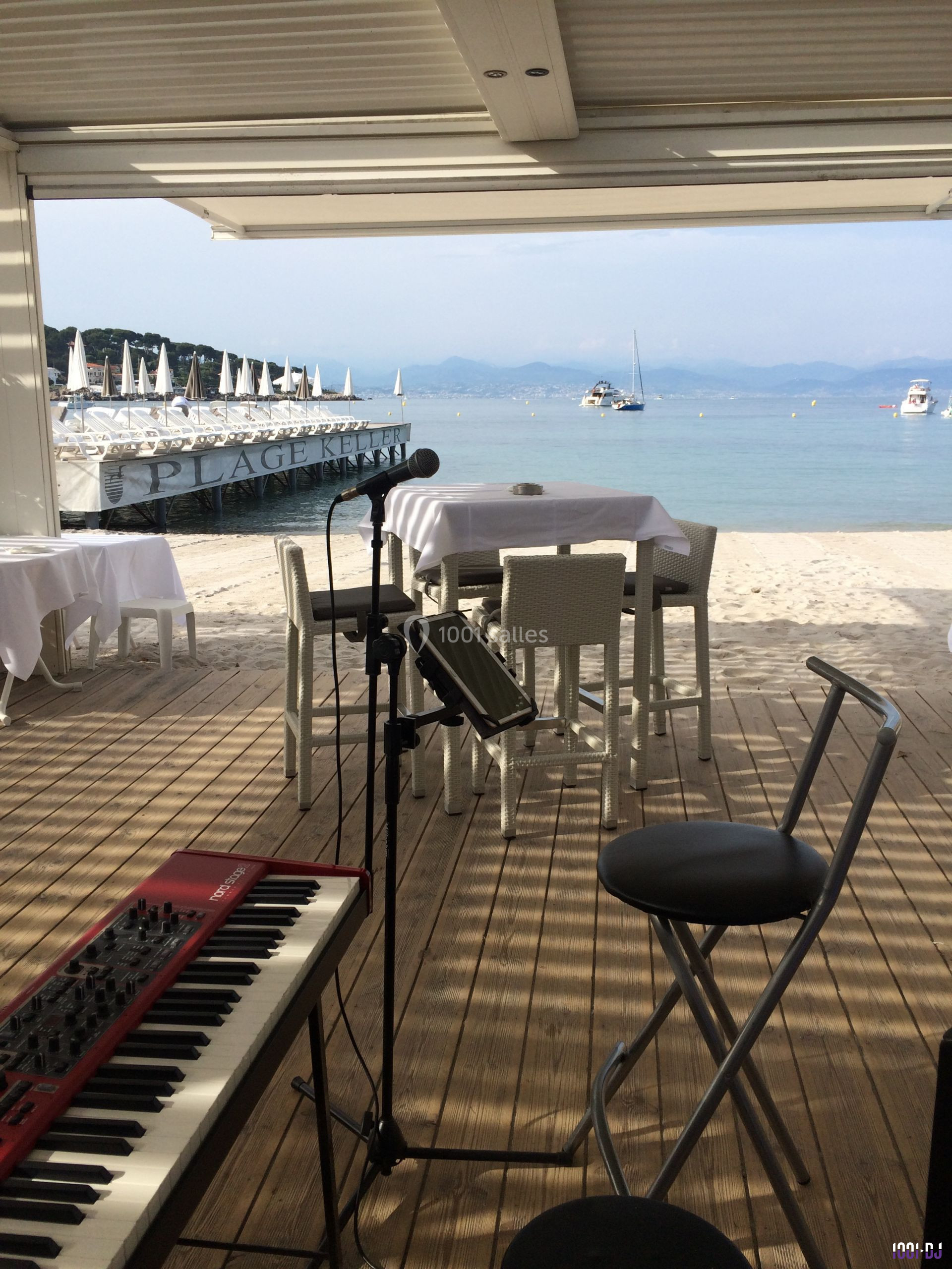 Vue d'une scène en bord de plage avec un clavier, un micro, des tables dressées et des bateaux au loin.