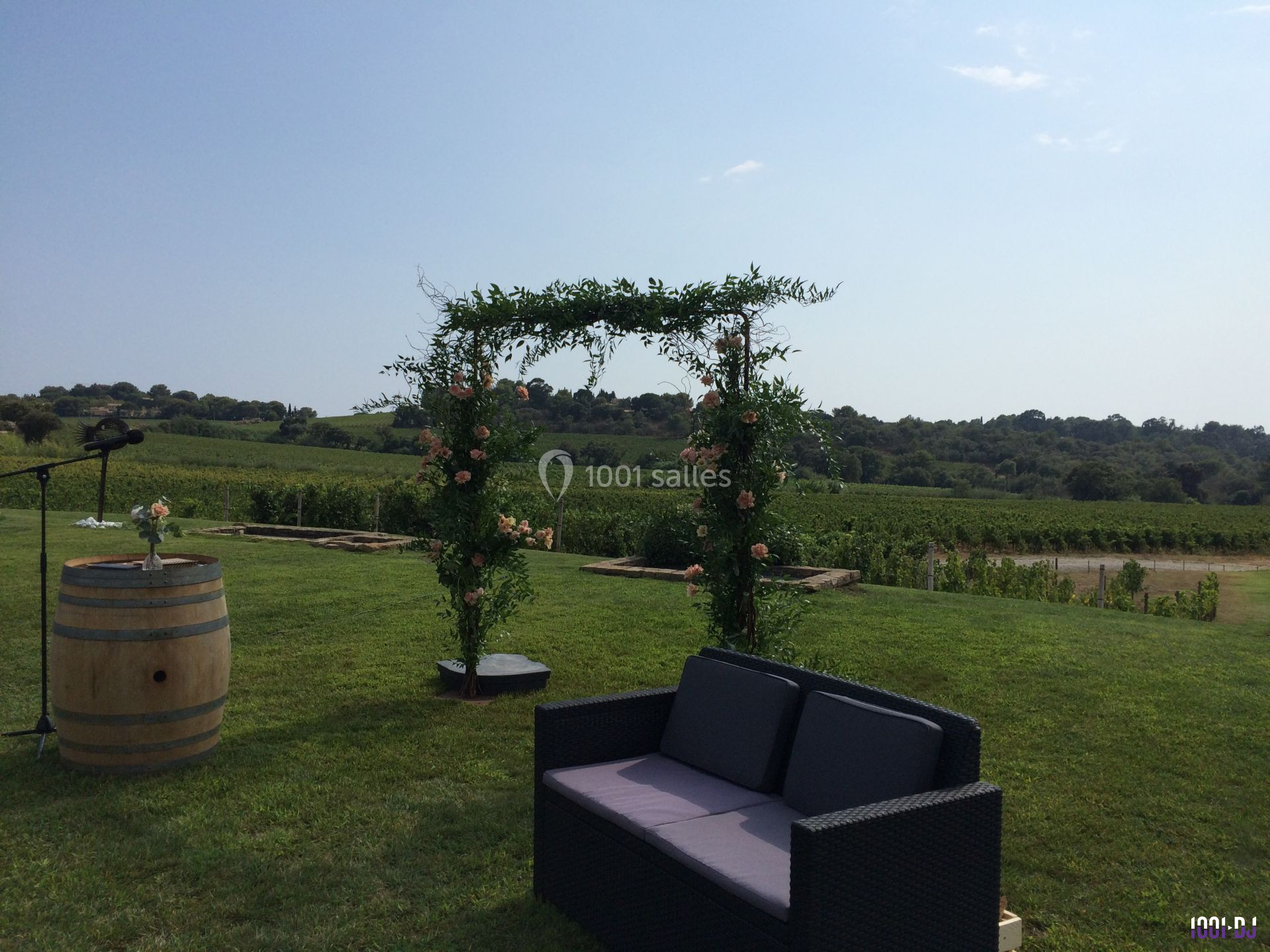 Arche fleurie, canapé en rotin et tonneau sur une pelouse avec vignoble en arrière-plan sous un ciel dégagé.