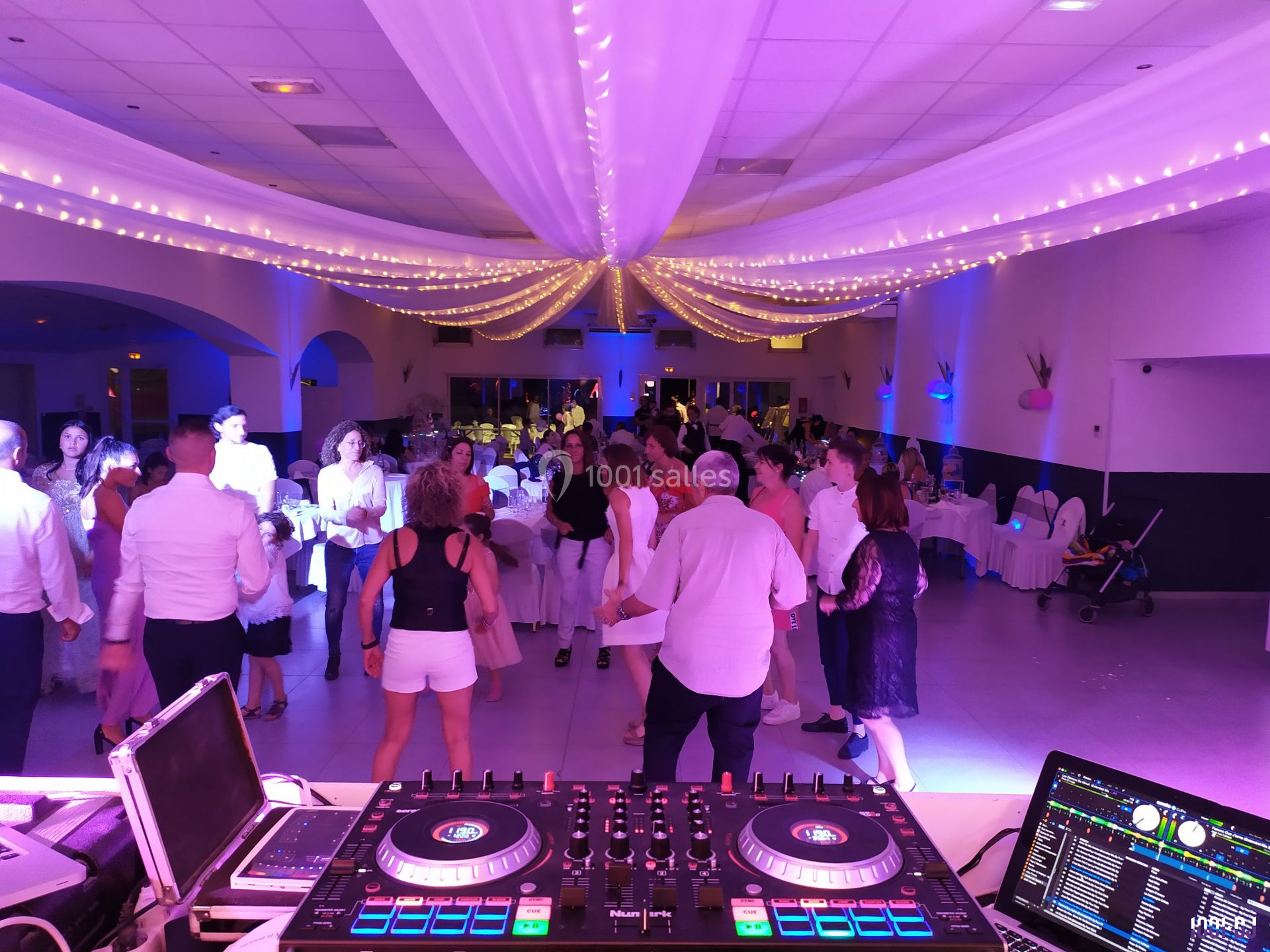 Des personnes dansent dans une salle décorée de guirlandes lumineuses, vue depuis une table de mixage de DJ.