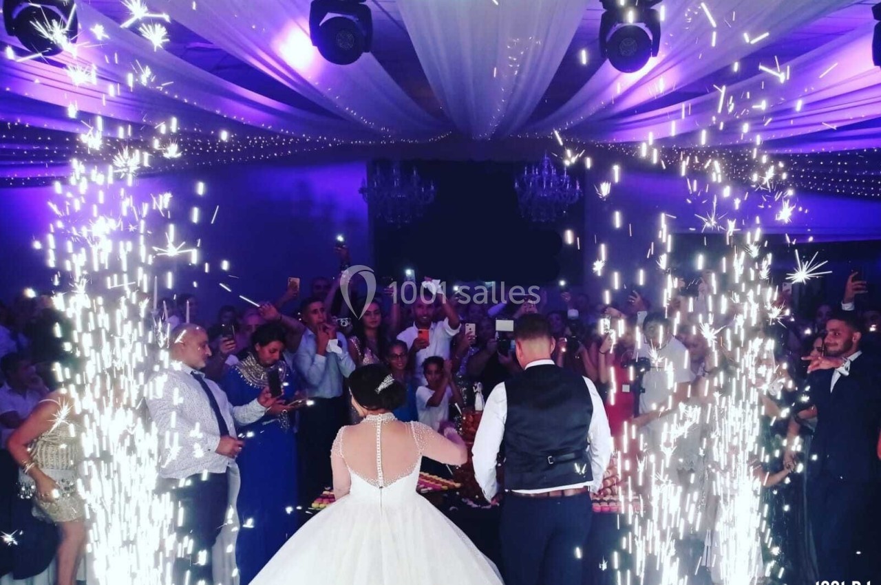 Un couple de mariés coupe leur gâteau entouré d'invités sous des étincelles et un éclairage violet festif.