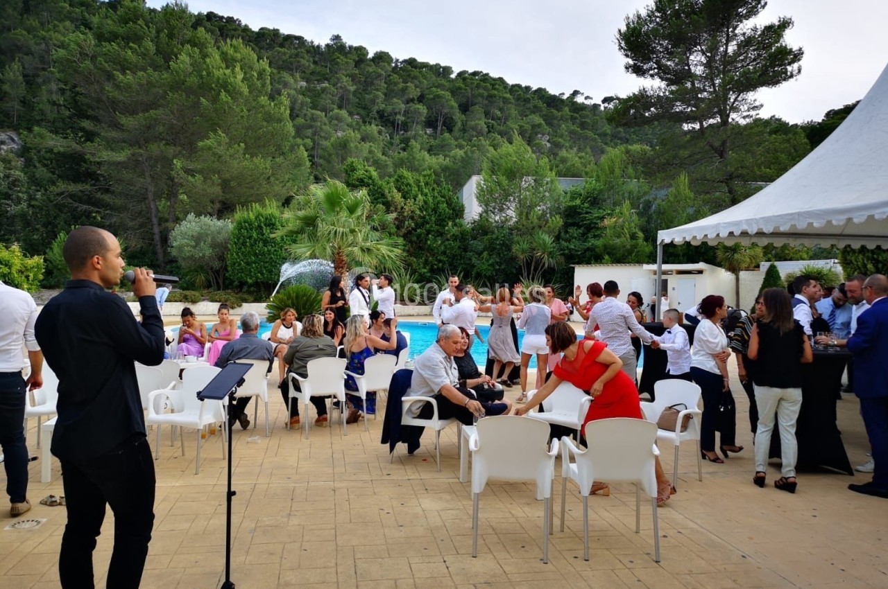 Groupe de personnes réunies en extérieur près d'une piscine, avec un chanteur et un micro au premier plan.
