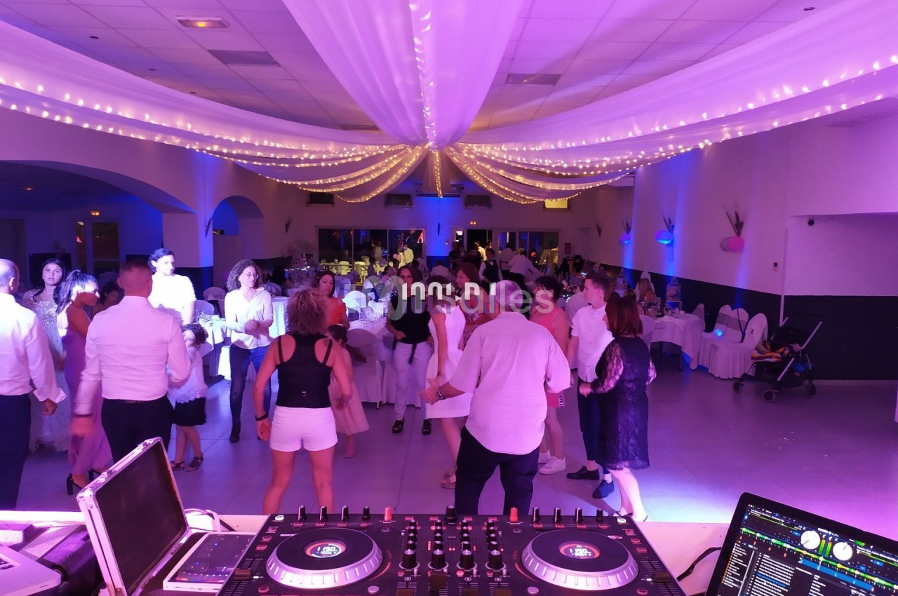 Salle de réception éclairée avec des guirlandes lumineuses, des invités dansant devant une table de mixage de DJ.