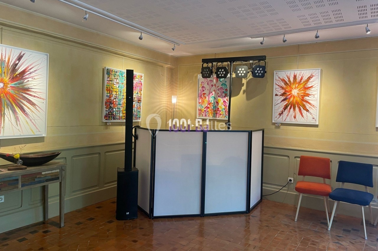 Salle d'exposition avec des peintures colorées accrochées aux murs, un pupitre et des chaises disposées autour.