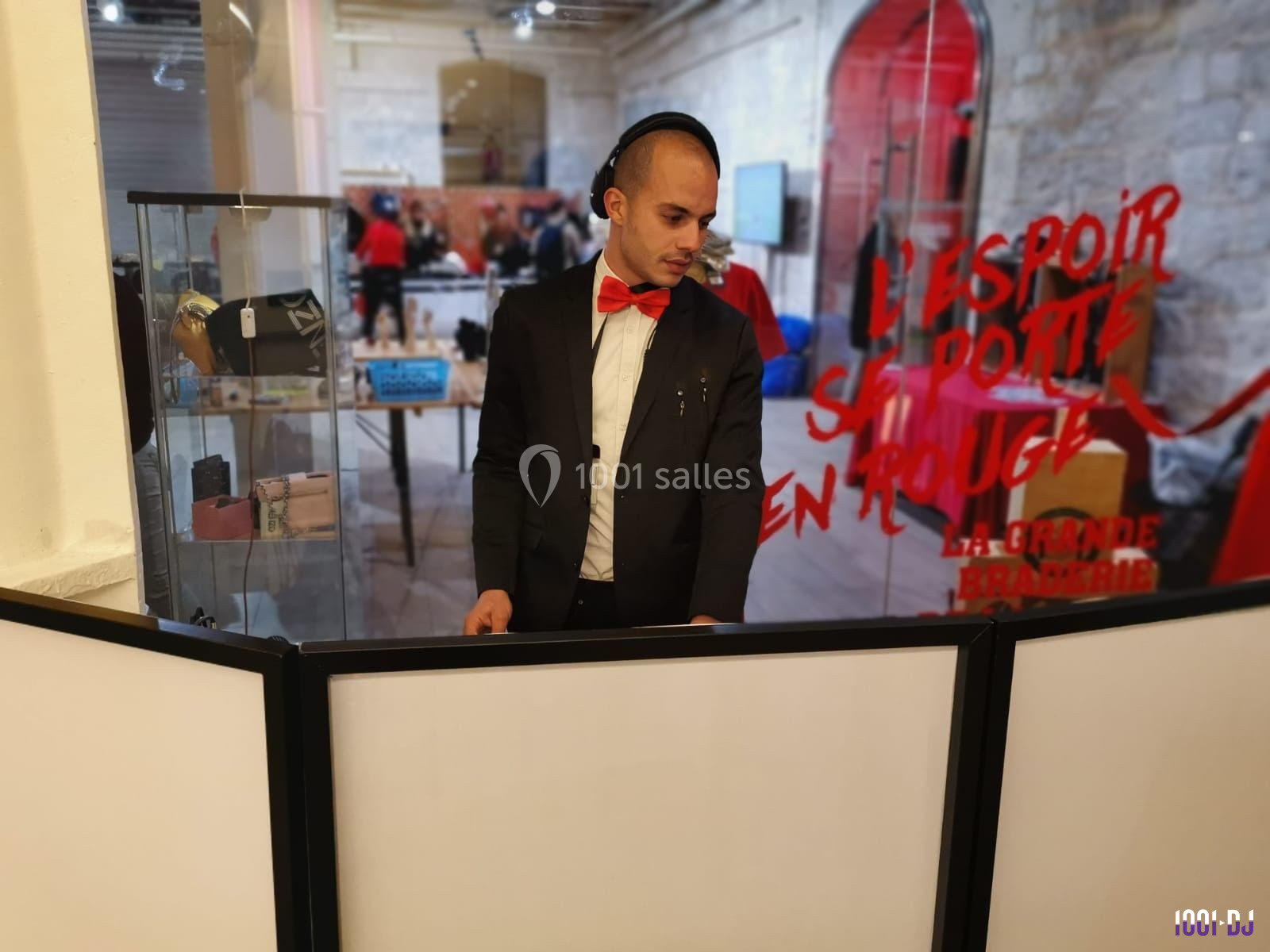 Un DJ en costume noir et nœud papillon rouge mixe devant un stand dans un espace intérieur.