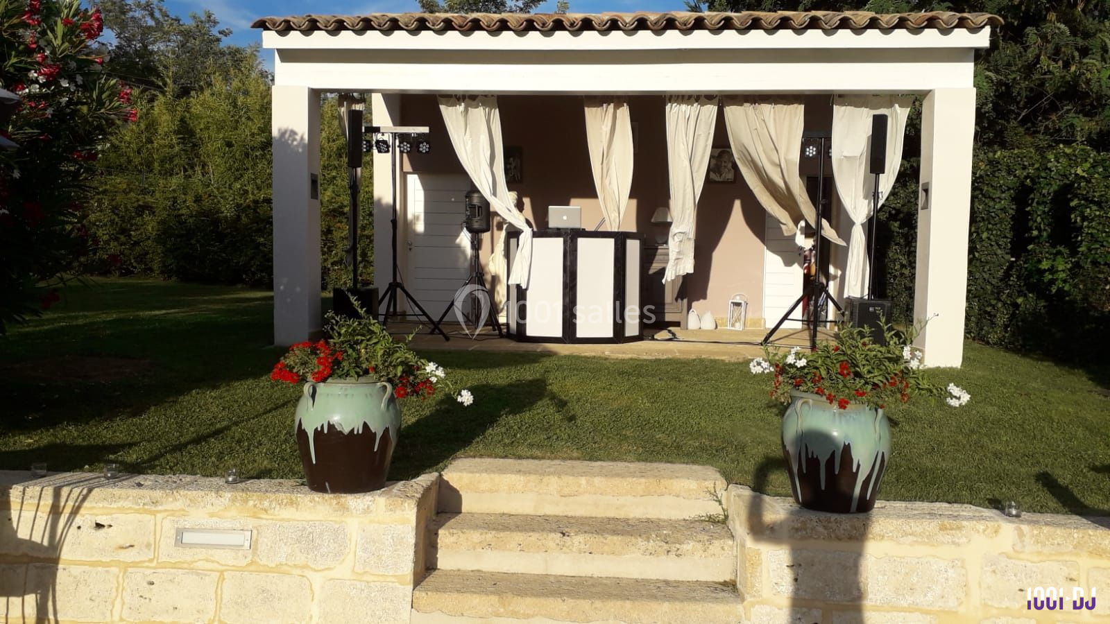 Pergola blanche avec rideaux ouverts, abritant une installation DJ, entourée de verdure et de pots de fleurs.