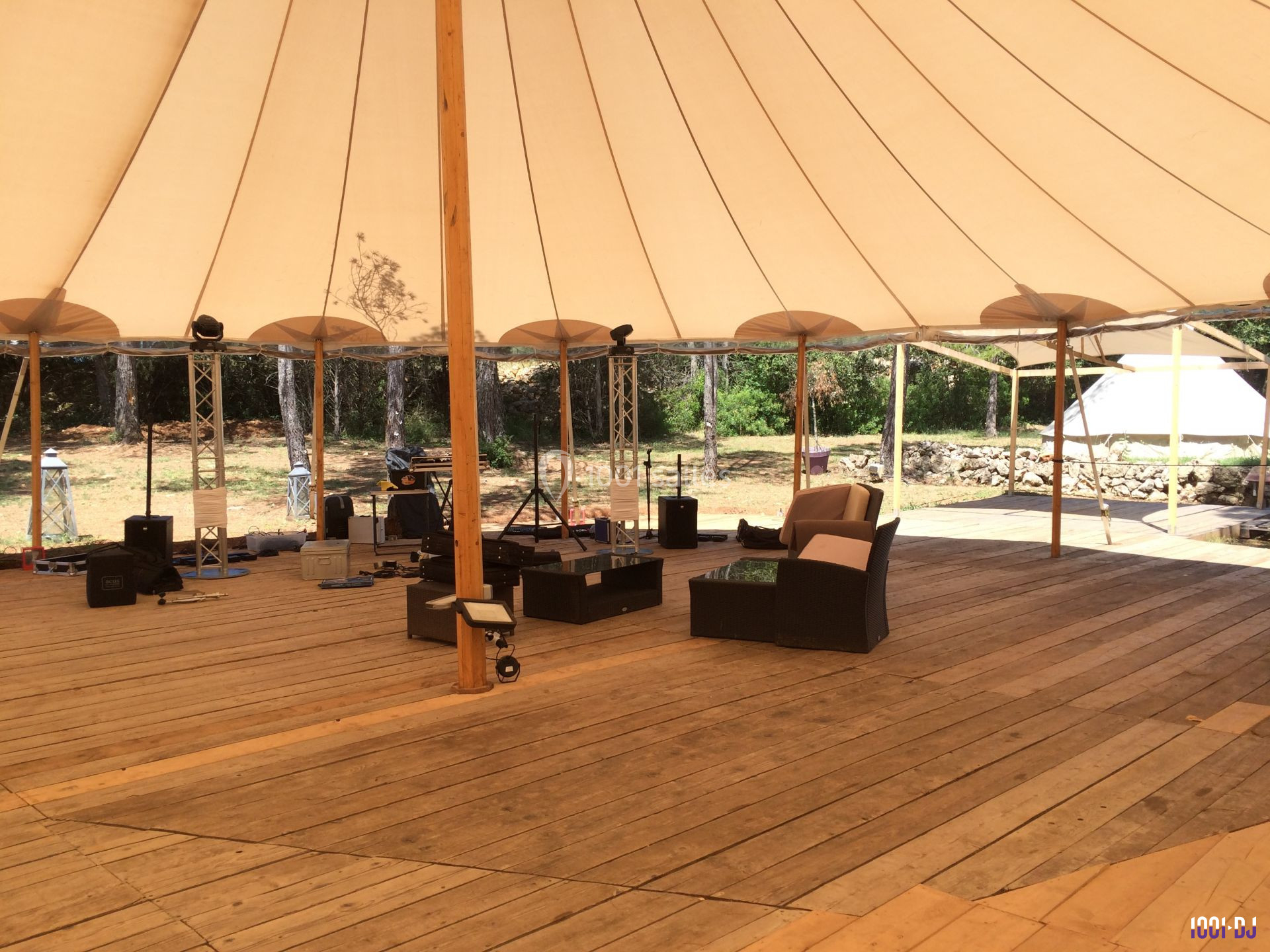 Intérieur d'une tente avec plancher en bois, mobilier d'extérieur et matériel technique, entourée de végétation.