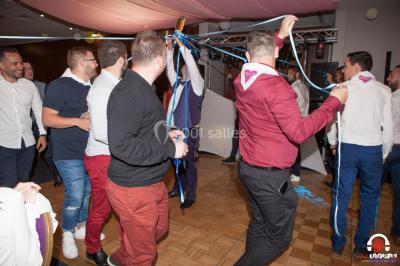 Des personnes dansent en ligne dans une salle décorée de ballons, avec des tables dressées et des convives en arrière-plan.