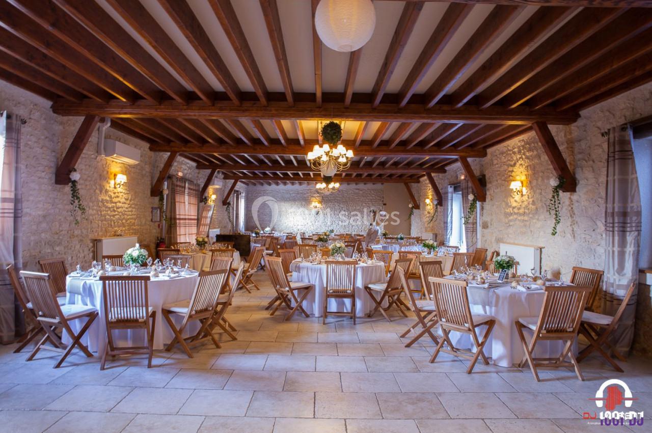 Salle de réception en pierre avec tables rondes dressées, chaises en bois et décoration florale.
