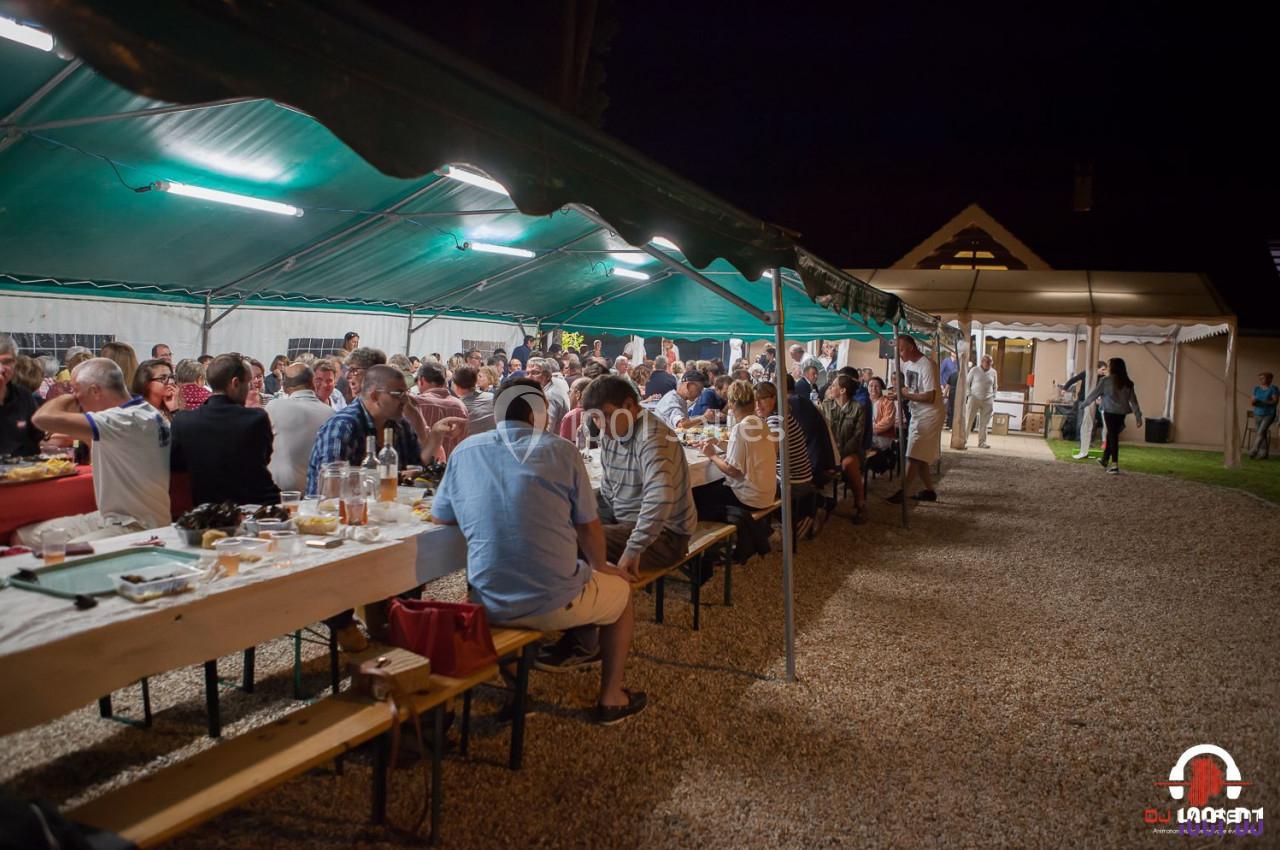 Groupe de personnes dînant sous une tente éclairée en soirée, avec des tables alignées sur un sol en gravier.