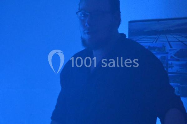 Homme barbu portant des lunettes dans une pièce éclairée par une lumière bleue, avec un tableau en arrière-plan.