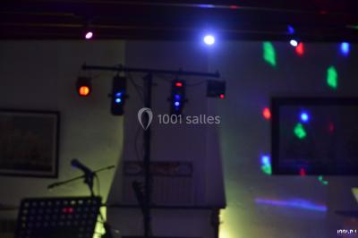 Salle polyvalente avec scène surélevée, rideaux bleus, écran blanc, tables et chaises disposées en avant-plan.