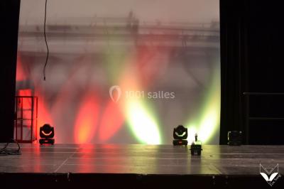 Salle polyvalente avec scène surélevée, rideaux bleus, écran blanc, tables et chaises disposées en avant-plan.