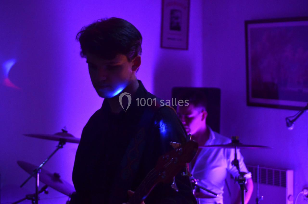 Un musicien joue de la guitare basse sous une lumière violette, avec un batteur en arrière-plan.