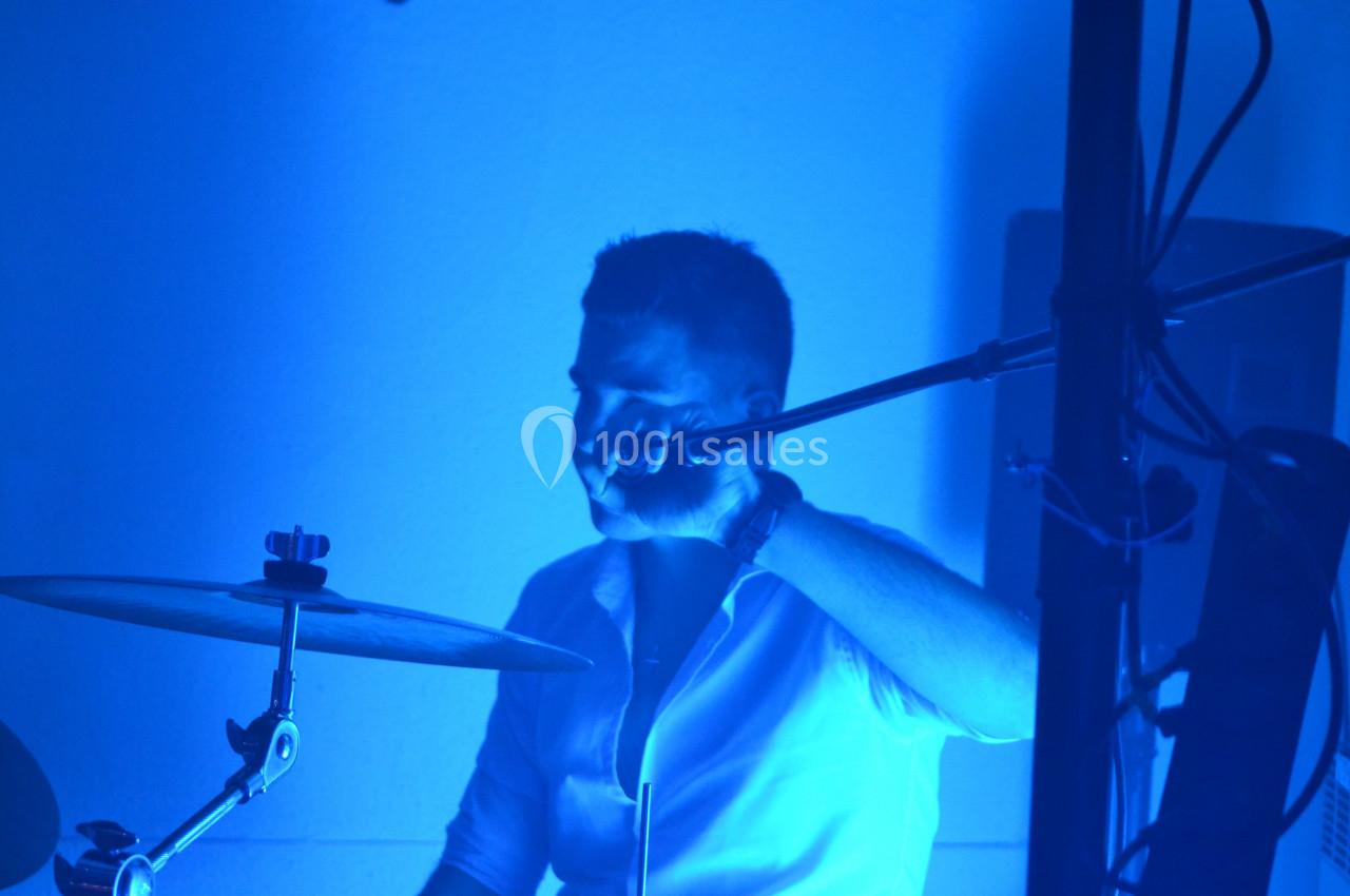 Un batteur en chemise blanche joue sur une batterie sous un éclairage bleu intense.