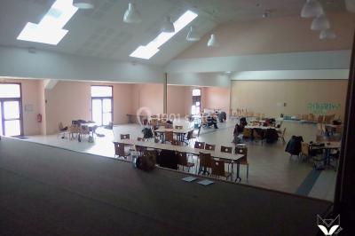 Salle polyvalente avec scène surélevée, rideaux bleus, écran blanc, tables et chaises disposées en avant-plan.