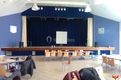 Salle polyvalente avec scène surélevée, rideaux bleus, écran blanc, tables et chaises disposées en avant-plan.