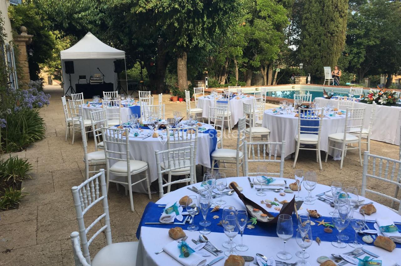 Tables rondes dressées pour un événement en plein air, avec nappes blanches, décorations bleues et une piscine en arrière…