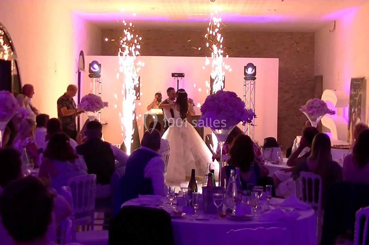 Un couple danse au centre d'une salle de réception décorée, entouré d'invités assis et d'effets pyrotechniques.