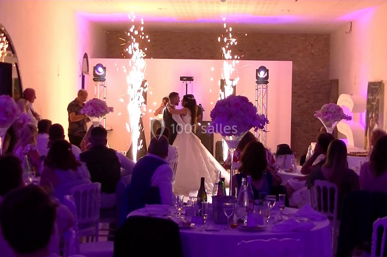 Un couple danse au centre d'une salle de réception éclairée en violet, entouré d'invités assis et de jets d'étincelles.