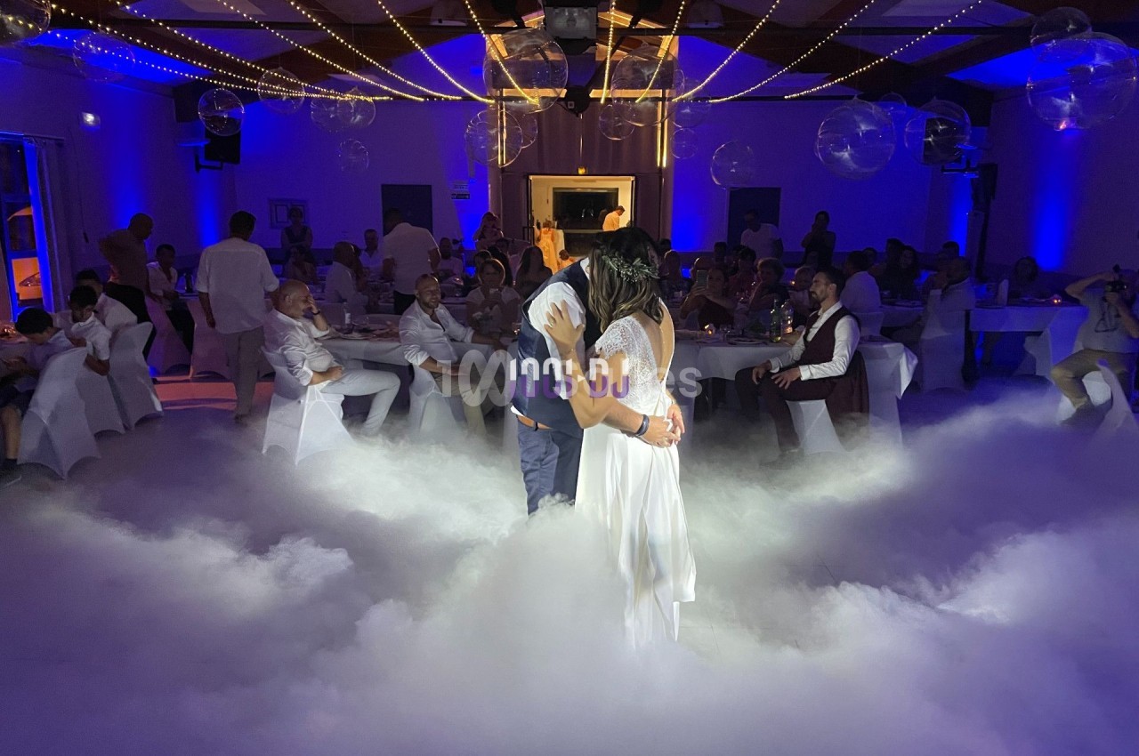 Un couple danse sur une piste enveloppée de fumée, entouré d'invités assis dans une salle illuminée.