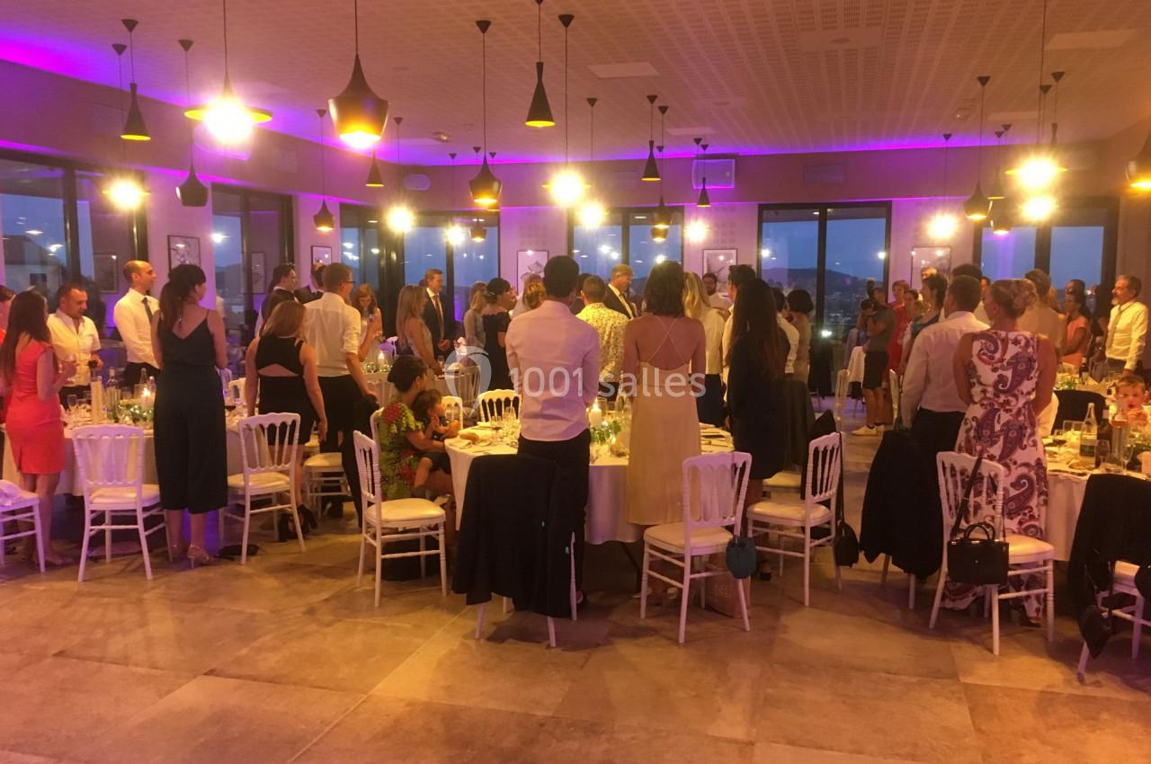 Salle de réception éclairée avec des invités debout autour de tables décorées pour un événement festif.