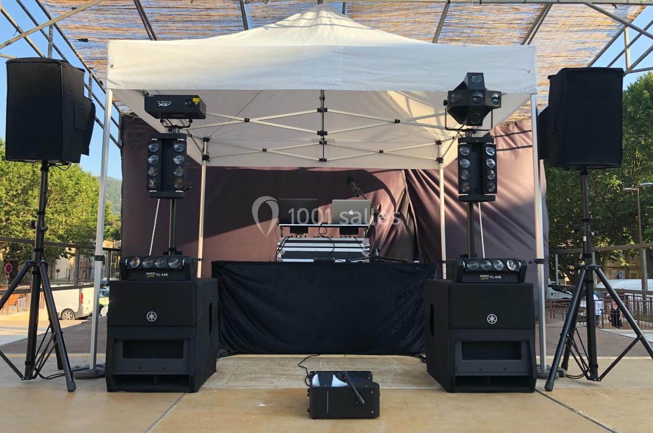 Scène extérieure avec équipement de sonorisation sous une tente blanche, incluant haut-parleurs et matériel DJ.