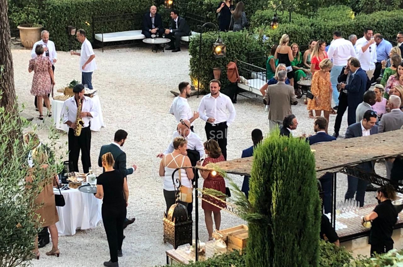 Groupe de personnes rassemblées dans un jardin pour un événement, avec buffet et musiciens en arrière-plan.