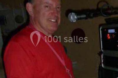 Homme souriant debout devant un micro, portant une chemise noire avec un gilet rayé, fond marron.