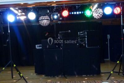 Homme souriant debout devant un micro, portant une chemise noire avec un gilet rayé, fond marron.