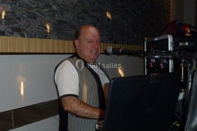 Homme souriant debout devant un micro, portant une chemise noire avec un gilet rayé, fond marron.