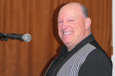 Homme souriant debout devant un micro, portant une chemise noire avec un gilet rayé, fond marron.