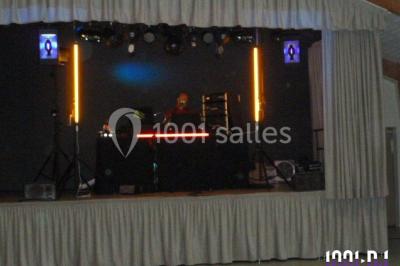 Homme souriant debout devant un micro, portant une chemise noire avec un gilet rayé, fond marron.