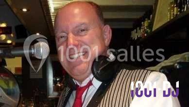 Homme souriant debout devant un micro, portant une chemise noire avec un gilet rayé, fond marron.