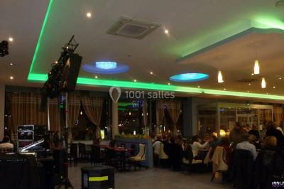 Homme souriant debout devant un micro, portant une chemise noire avec un gilet rayé, fond marron.