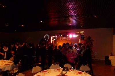 Homme souriant debout devant un micro, portant une chemise noire avec un gilet rayé, fond marron.