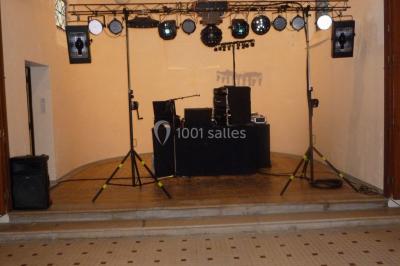 Homme souriant debout devant un micro, portant une chemise noire avec un gilet rayé, fond marron.