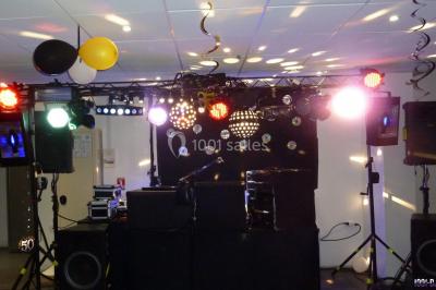 Homme souriant debout devant un micro, portant une chemise noire avec un gilet rayé, fond marron.