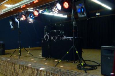 Homme souriant debout devant un micro, portant une chemise noire avec un gilet rayé, fond marron.