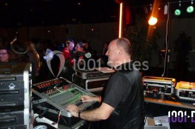 Homme souriant debout devant un micro, portant une chemise noire avec un gilet rayé, fond marron.