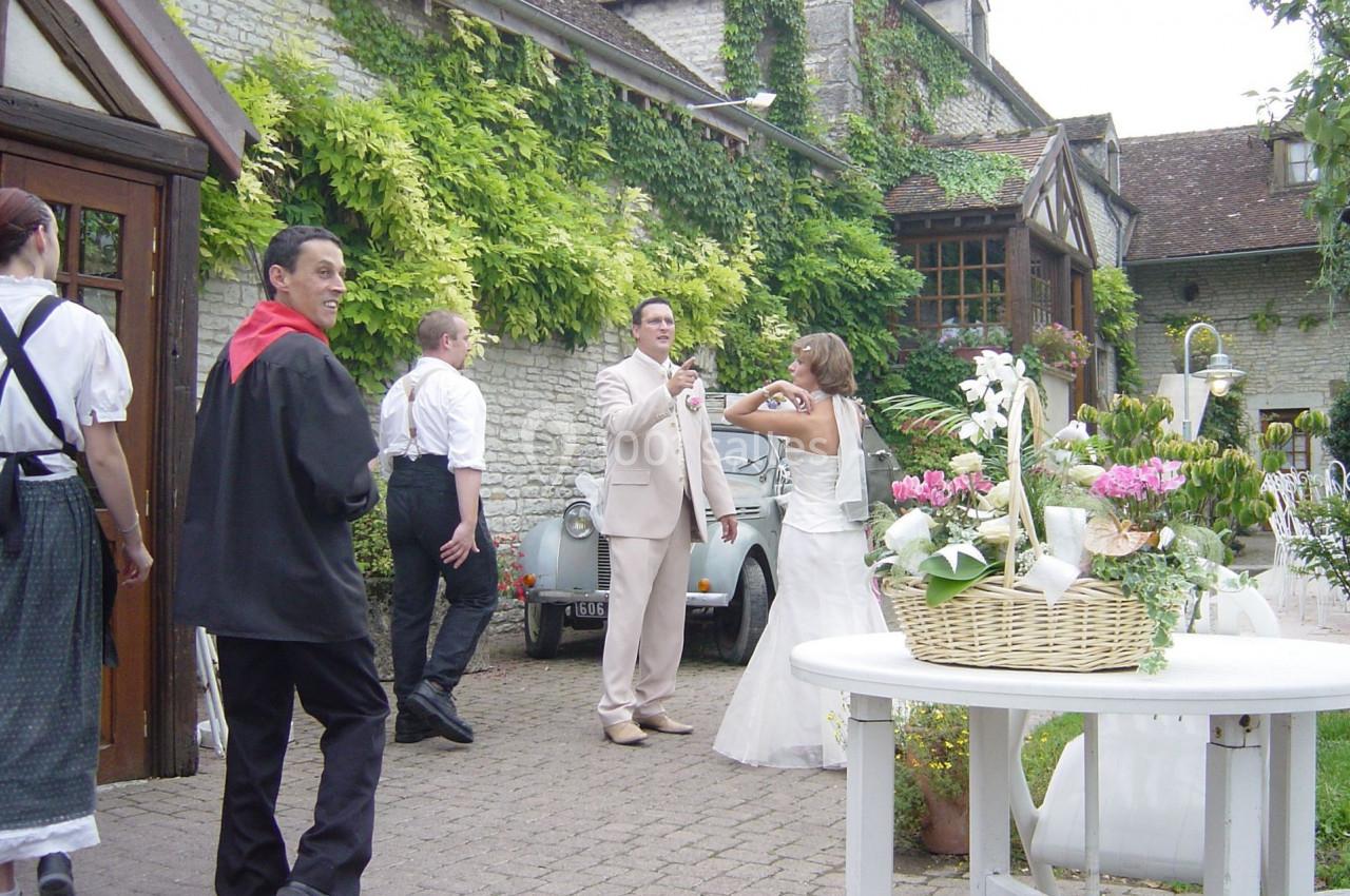 Un couple en tenue de mariage pose devant une voiture ancienne, entouré d'invités dans une cour fleurie.