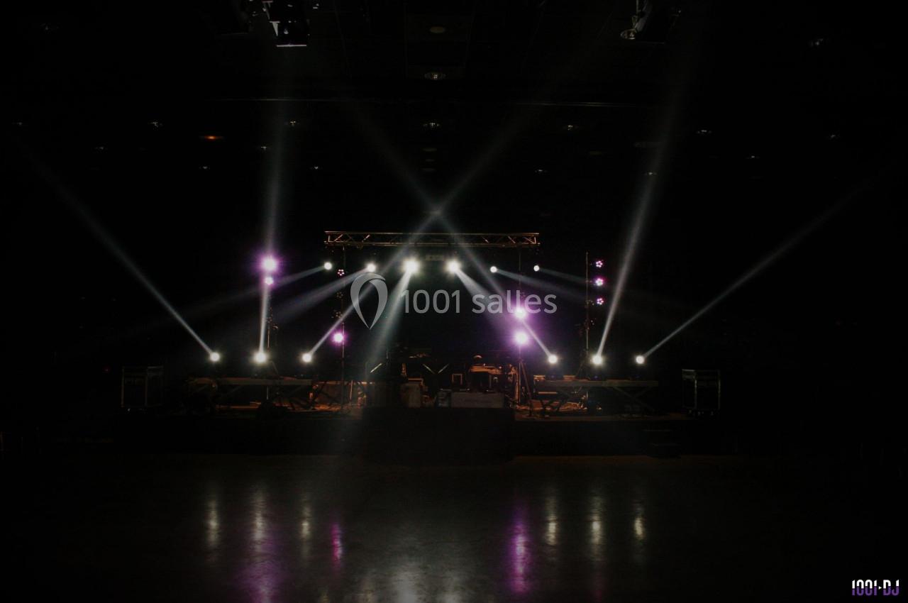 Scène de concert vide éclairée par des projecteurs, avec des équipements et instruments visibles dans une salle sombre.