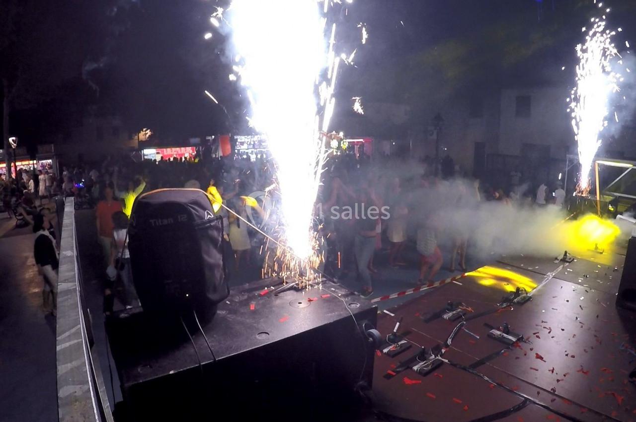 Scène de concert en plein air avec des effets pyrotechniques et une foule dansant sous des lumières colorées.