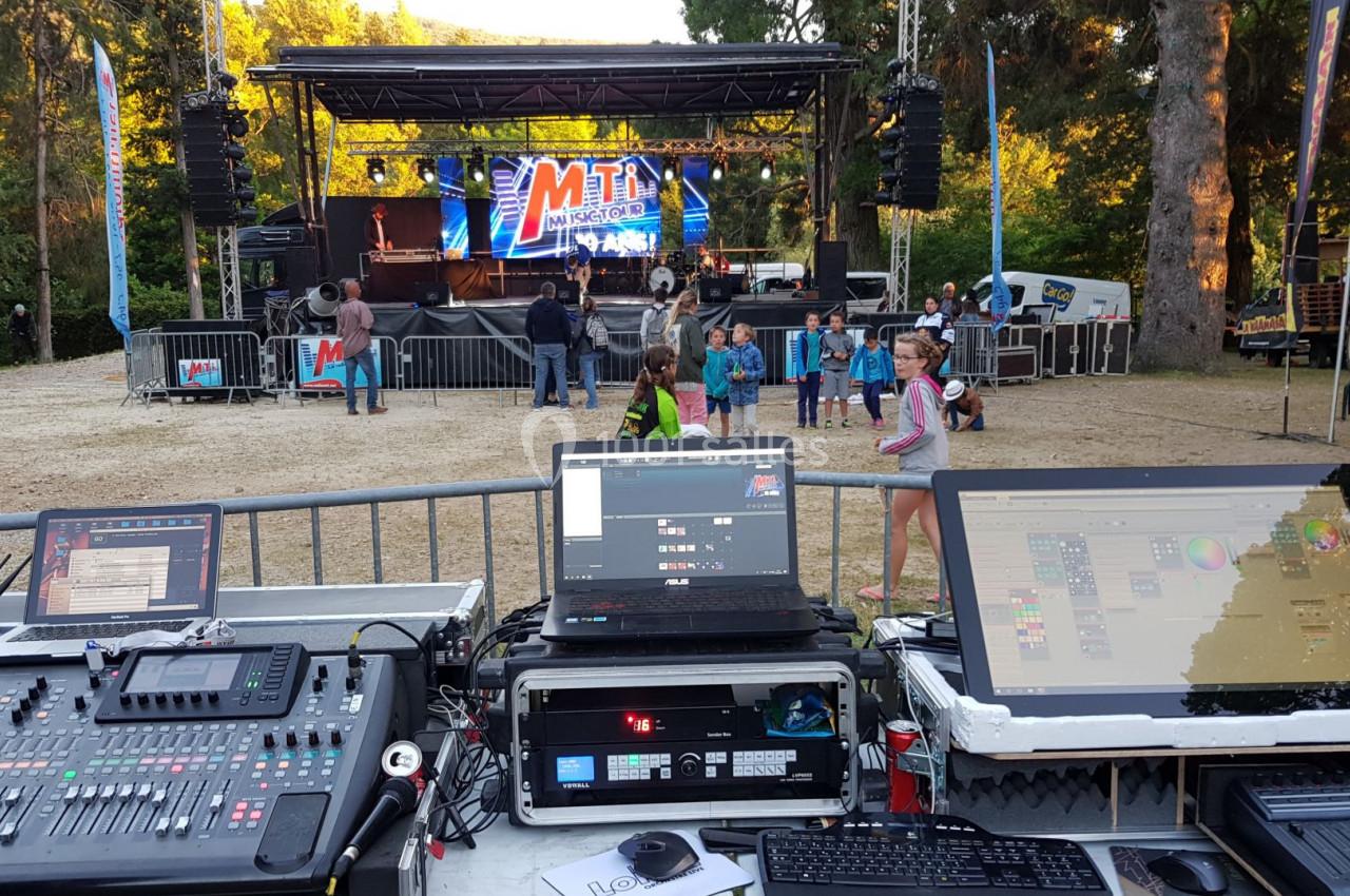 Poste de régie technique avec consoles et écrans, face à une scène extérieure en préparation pour un événement.