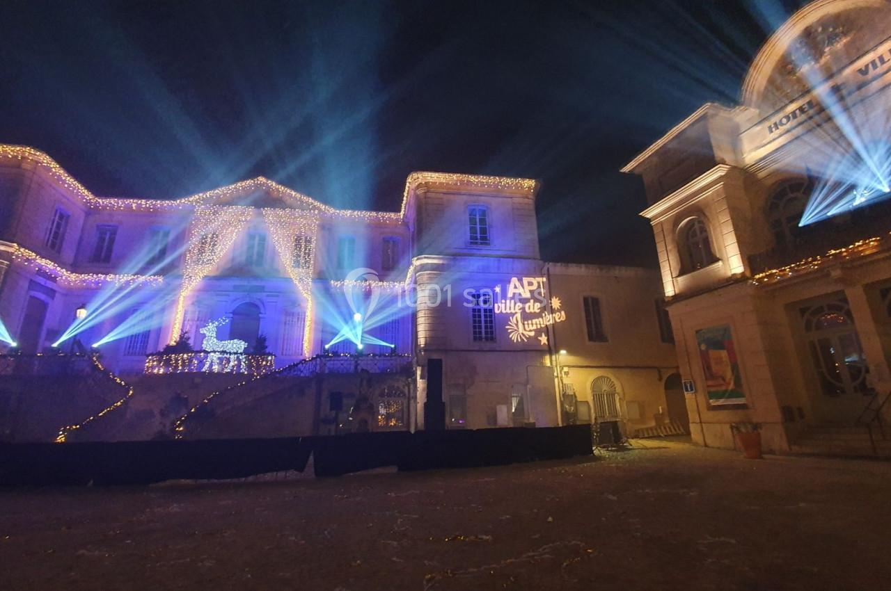 Façade illuminée d'un bâtiment avec décorations de Noël, projecteurs colorés et inscription ’APT ville de lumière’.